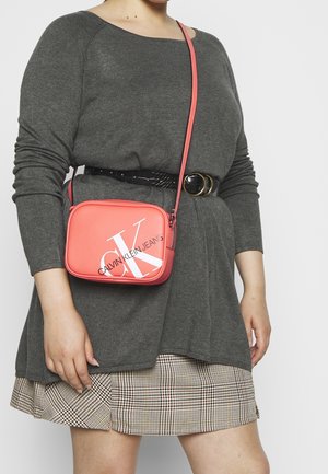 Koralowa torebka crossbody Calvin Klein Jeans z białym logo, prostokąt kształt, gładka faktura i regulowany pasek noszona na szarym topie i kratkowanej spódnicy.