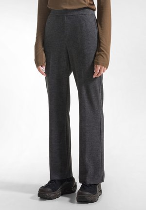 Persoon draagt donkergrijze geribde rechte broek, zwarte chunky sneakers en een lange mouw bruine top, staand op een witte achtergrond.