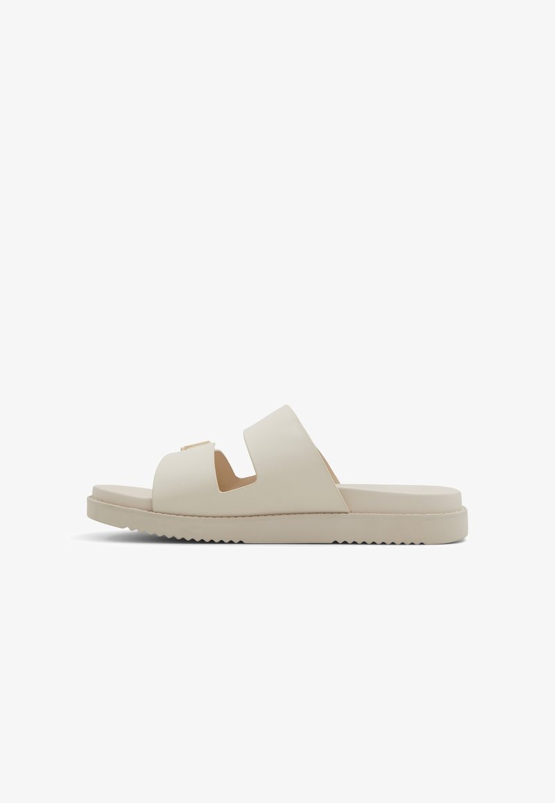 Beige slide sandal with dual cut-out straps, flat sole, and textured bottom. Made of smooth material, featuring minimalistic design accents.