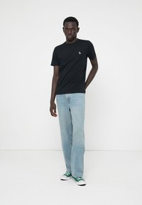 Black short-sleeve T-shirt with a small logo on the chest, paired with light blue wide-leg jeans and green-accented sneakers.