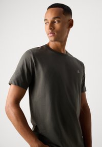 Young man with short hair wearing a dark olive green T-shirt, looking to his left against a plain light background.