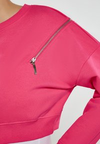 Ružová mikina s štruktúrovaným povrchom, okrúhlym výstrihom a dlhými rukávmi. Výrazný strieborný zips v hornej ľavej časti pre ďalší dizajnový prvok.