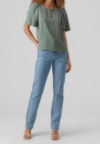 Blusa verde oliva con maniche a sbuffo e dettaglio a forma di chiave sul collo, abbinata a jeans azzurri a gamba dritta e sandali beige.