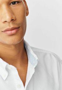 Camisa blanca de botones con un cuello interior a rayas azules, que presenta una textura suave y un corte relajado, destacando el detalle del cuello cerca del cuello.
