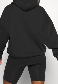 Haut à capuche noir surdimensionné avec une capuche, en tissu texturé et épaules tombantes, associé à un short noir ajusté, tous deux dans une couleur unie.