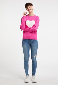 Pulover roz cu un mare design de inimă albă cu nervuri, asortat cu blugi skinny decolorati, de un albastru deschis și adidași albi.