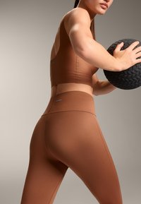 Barna, aktív sportmelltartó és leggings, sima anyagból készült, illeszkedő kialakítással, minimális varrásokkal és diszkrét márka logóval díszítve.