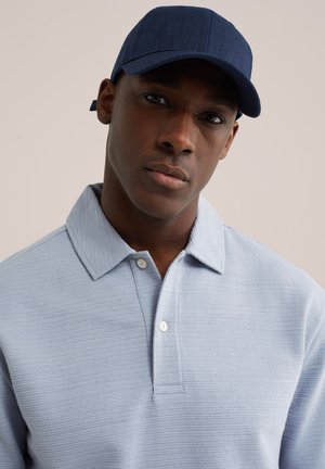 Homme portant une casquette marine et un polo bleu clair texturé, face à l'avant avec une expression neutre sur un fond uni.