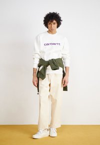 Sweatshirt blanc avec logo "carhartt." violet, pantalon crème large et baskets blanches, avec une veste verte nouée à la taille.