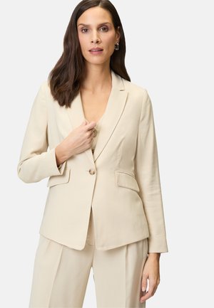 Veste tailleur beige en tissu doux, dotée d'une fermeture à un bouton, d'un col en cranté et de deux poches avant. Texture lisse.