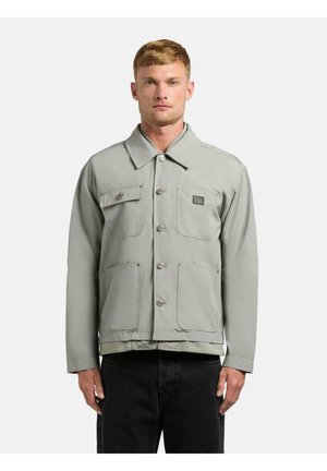 Mężczyzna ubrany w jasnoszarą kurtkę na guziki z czterema przednimi kieszeniami, stojący na tle jednolitego, białego tła.