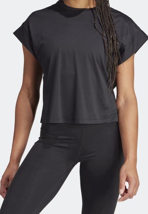 Frau trägt ein schwarzes kurzärmeliges, geripptes Crop-Top und schwarze Leggings, steht mit einer Hand entspannt an der Seite.
