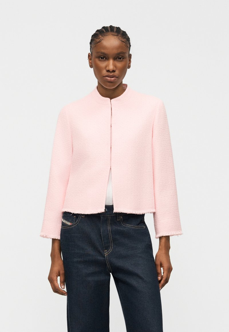 Γυναίκα με πλεξούδες φοράει ανοιχτό ροζ μακρυμάνικο σακάκι με υφή πάνω από λευκό τοπ και σκούρο μπλε τζιν, στέκεται μπροστά σε απλό φόντο.