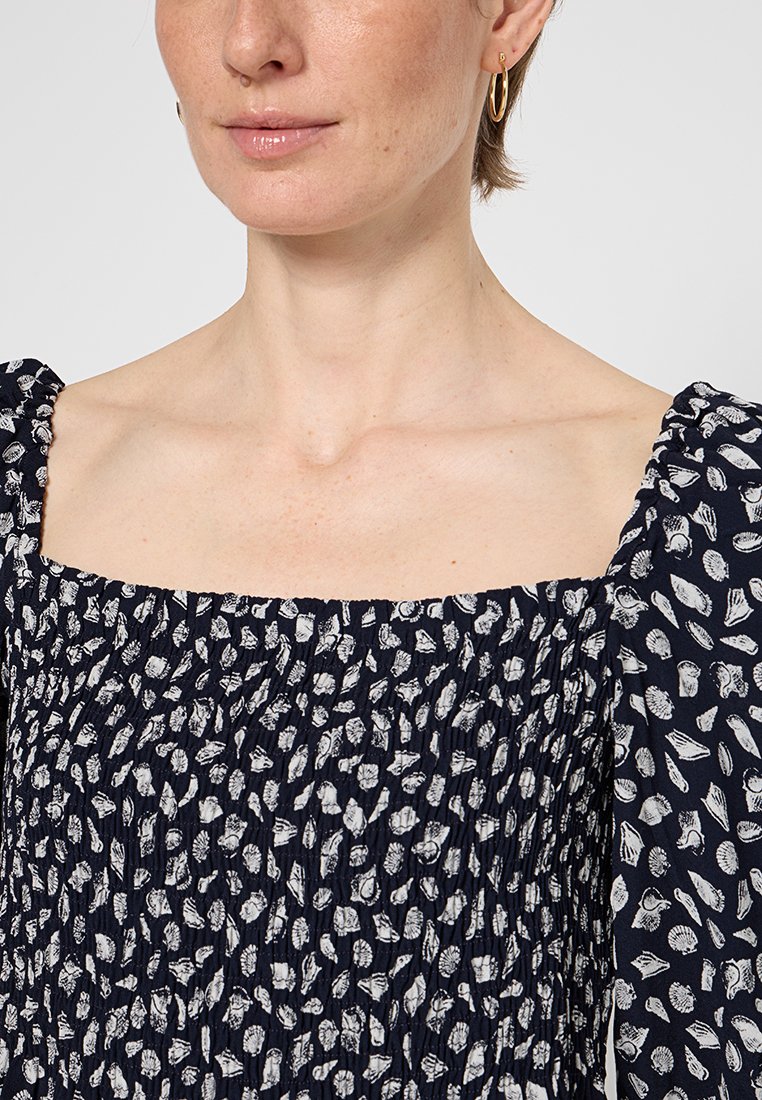 Frau trägt ein marineblaues, gerafftes Oberteil mit weißen Muschelmustern und goldenen Creolen, die Nacken und Schultern zeigend.