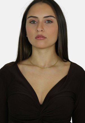 Joven con cabello largo y oscuro, vestida con una blusa negra de escote en V y un delicado collar dorado, mirando al frente con expresión neutral.