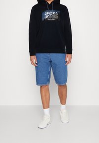 Černá mikina s kapucí a potiskem loga, v kombinaci s modrými džínovými šortkami. Bílé tenisky s šedými detaily a bílé ponožky, stojící na neutrálním pozadí.