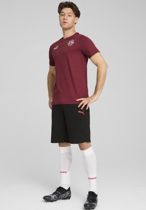 Bordeaux kortærmet t-shirt med et subtilt mønster og logo, parret med sorte shorts med et rødt mærkelogo, hvide sokker og sorte fodboldstøvler.