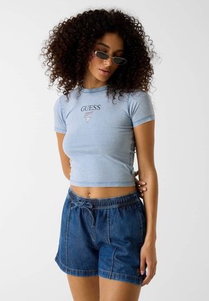 Giovane donna con capelli ricci che indossa piccoli occhiali da sole, una t-shirt corta Guess azzurra chiaro e pantaloncini di jeans su sfondo neutro.