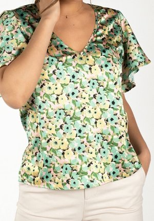 Kvinde iført kortærmet V-hals bluse med grønt, gult og blåt blomsterprint, kombineret med lyse beige bukser.