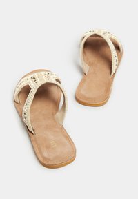 Sandali piatti con perline, due cinturini per le dita, una soletta in simil suede beige e una suola liscia color tan. Accentuati da dettagli metallici e testurizzati.