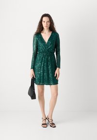 Abito verde con paillettes, con maniche lunghe, scollatura a V profonda, vita aderente e orlo corto. Accostato a sandali neri con cinturini e una piccola borsa.