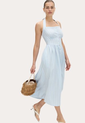 Vrouw draagt een lichtblauwe gestreepte halter midi-jurk, houdt een bruine bucketbag met franjes vast en draagt witte sandalen met hak.