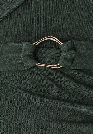 Grønt tekstureret stof med en knyttet strop og en cirkulær ring i guldfarvet metal som detalje. Materialet har en subtil glans og blødhed.