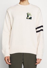 Krämsfärgad stickad tröja med rund halsringning, med en grön och gul logotypapplikation och marinblå ränder på ärmen.