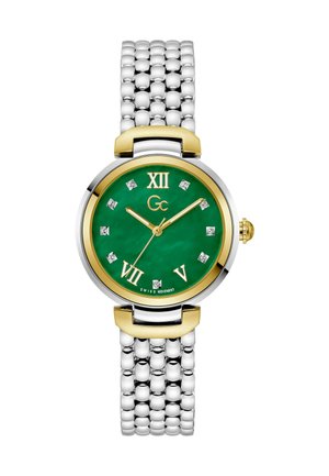 Ümar kell, millel on roheline emberdial, kuld- ja hõbedased aktsendid, rooma numbrid, kristallist tunni märgid ja hõbedane metallist rihm.