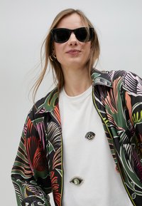 Šarena, uzorkovana jakna na munju u zelenim, ružičastim i crnim nijansama; bijela majica s tri pribadače u obliku oka; model nosi crne sunčane naočale.