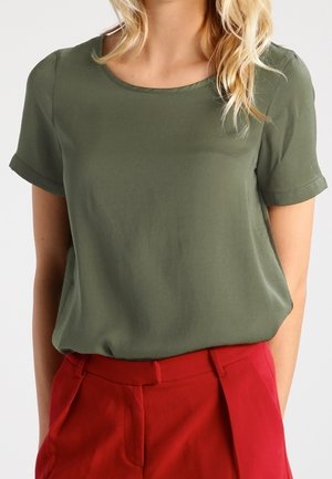 Bluzka z krótkim rękawem w kolorze oliwkowej zieleni, z okrągłym dekoltem, wykonana z gładkiego materiału, zestawiona z wysokim stanem czerwonymi spodniami.