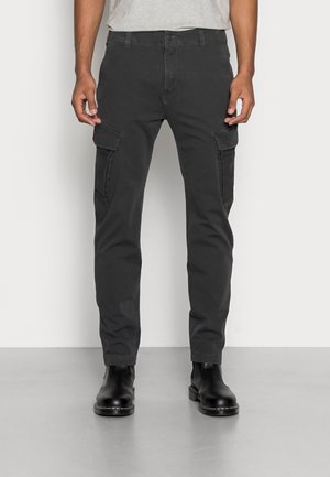 Man wearing black cargo pants with side pockets and black leather boots, standing on gray floor against white wall.