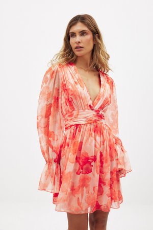 Kwiatowa mini sukienka w jasnym brzoskwiniowym kolorze z czerwonymi abstrakcyjnymi wzorami, głębokim dekoltem w kształcie litery V, plisowanym pasem i bufkami. Przezroczysty, lekki materiał.