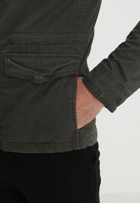 Grøn jakke med mat finish, der har en brystlomme, sidelommer og en slank pasform. Stoffet ser blødt og let ud.