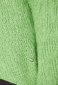 Tissu de pull doux en maille vert clair avec un petit emblème de marque en métal argenté rond près du bord inférieur.