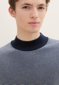 Gebreide pullover met een marineblauwe geribde kraag, met een textuurpatroon van witte stippen op een donkerblauwe achtergrond. Soepele stoftextuur.