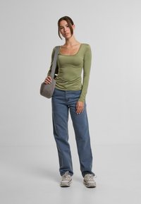 Langärmliges, olivgrünes Oberteil mit quadratischem Ausschnitt und gerafften Details, kombiniert mit hellblauen, geraden Jeans und grauer Umhängetasche.