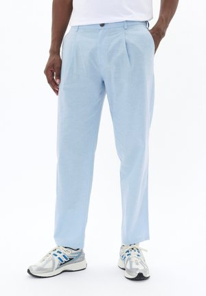 Mann trägt hellblaue, plissierte Hose und weiße Sportschuhe mit blauen und grauen Akzenten, steht mit entspannten Händen.