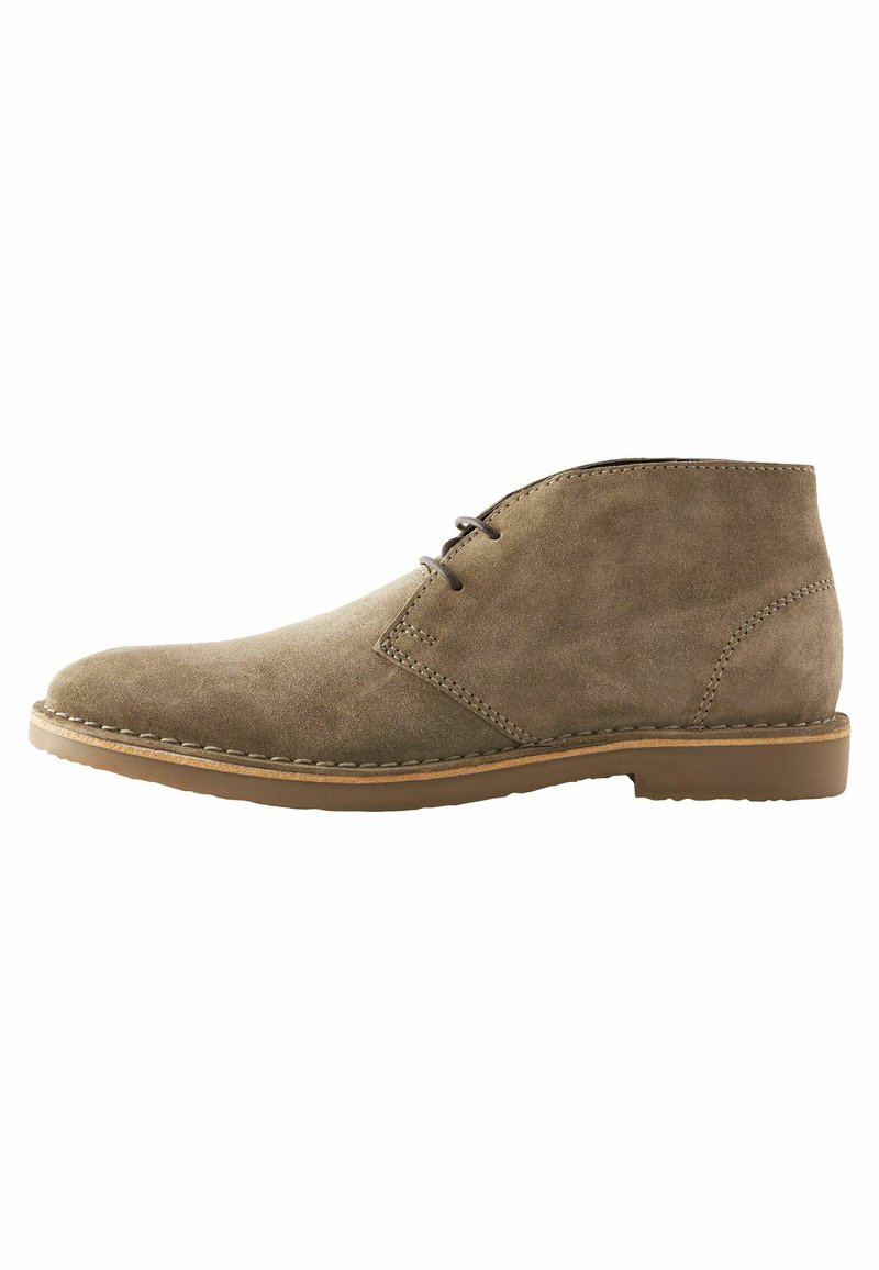 Botas de ante marrón hasta el tobillo con punta redondeada, dos ojetes para los cordones y una suela de goma color beige. Detalles de costura visibles cerca de la suela.