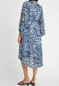 Blå och svart mönstrad klänning med långa ärmar, cinchad midja och fladdrig kjol, tillverkad av lätt material. Sandaler synliga på fötterna.