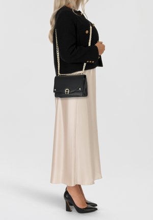 Giacca nera corta con bottoni dorati, abbinata a un abito lungo di raso beige. Borsa nera in pelle con tracolla a catena dorata e dettaglio di chiusura.