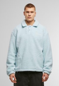 Világoskék fleece pulóver, galléros nyakkal, gombos záródással, és hímzett logóval a bal mellrészen. Oldalsó zsebekkel rendelkezik.