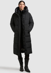 Un manteau matelassé noir présente un design long et rembourré avec des fermetures éclair et des boutons-pression, une grande capuche et des poches avant. Porté sur une tenue sombre.