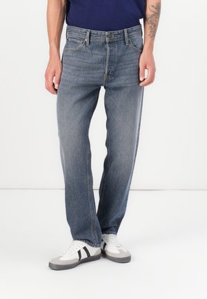 Homme portant un jean bleu délavé et des baskets blanches avec des rayures noires sur fond blanc, avec un tatouage visible sur l'avant-bras gauche.