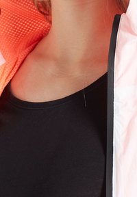 Czarny top na ramiączkach pod rozpiętą jasnoróżową i pomarańczową kurtką z teksturowanym wnętrzem. Tkanina wydaje się gładka i dopasowana.