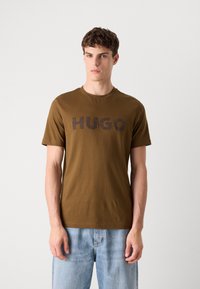 Barna pamut póló rövid ujjakkal és kerek nyakkivágással, amelyen egy nagy, texturált "HUGO" logó látható sötét színben a mellkason.