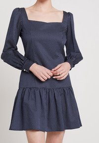 Vestido azul marino con lunares, escote cuadrado, mangas largas abullonadas, cuerpo ajustado y un dobladillo acampanado con volantes. Fabricado con una tela suave.