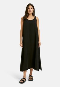 Schwarzes ärmelloses Maxikleid mit Rundhalsausschnitt und A-Linie. Der Stoff ist glatt mit einem leichten Glanz; getragen mit schwarzen Sandalen.