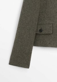 Manche gauche et partie inférieure d'un manteau en laine gris foncé avec poche à rabat boutonnée sur fond blanc.