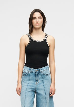 Femme portant un haut noir sans manches avec des clous argentés sur le col et les bretelles, associé à un jean taille haute bleu clair.